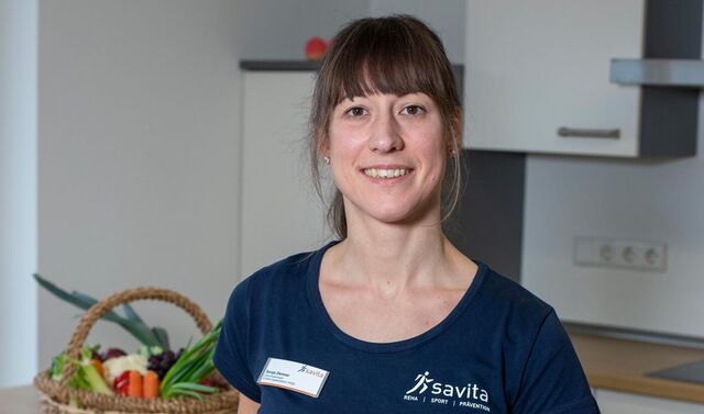 Sonja Denner steht vor der Lehrküche. Im Hintergrund ist ein gut gefüllter Obstkorb zu sehen.