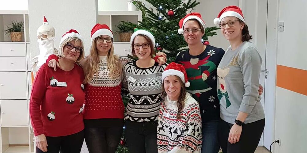 Sechs Mitarbeiterinnen mit Weihnachtsmützen posieren vor einem Weihnachtsbaum.