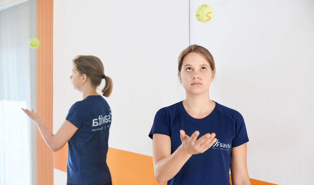 Ein junge Trainerin in blauem savita Shirt wirft einen Tennisball mit Buchstaben hoch. Sie steht vor einem Spiegel, der die Aktion gespiegelt abbildet.