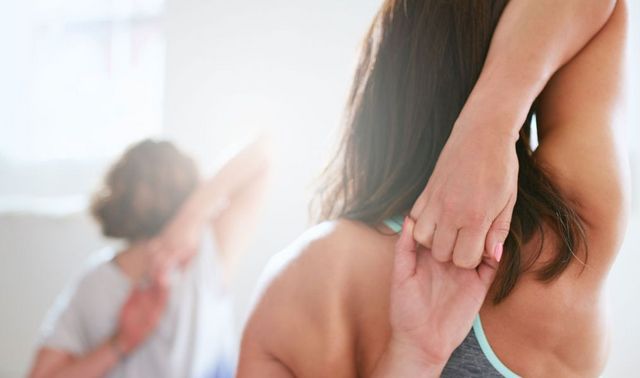 Die Oberkörper von zwei jungen Frauen sind von hinten zu sehen. Sie dehnen sich die Schultern, indem die Hände hinter dem Hals zusammenfassen.