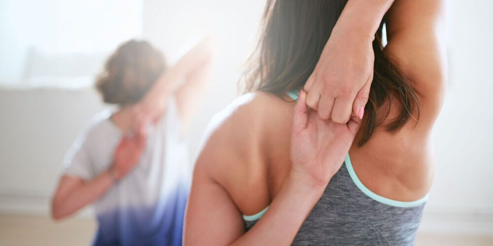 Die Oberkörper von zwei jungen Frauen sind von hinten zu sehen. Sie dehnen sich die Schultern, indem die Hände hinter dem Hals zusammenfassen.