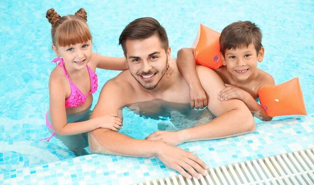 Ein junger Mann ist mit einem kleinen Jungen, welcher Schwimmflügel trägt und einem kleinem Mädchen im Schwimmbad am Beckenrad. Sie schauen alle drei lächelnd in die Kamera und halten sich am Beckenrad fest. 