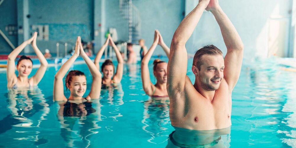 Ein Wassergymnastik-Kurs: Der muskulöse Kursleiter ist im Vordergrund zu sehen. Er streckt seine Arme in die Höhe. Dahinter machen vier Kursteilnehmerinnen die Übung nach.