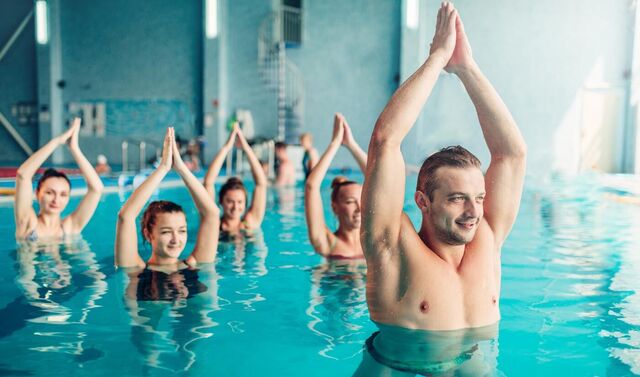 Ein Wassergymnastik-Kurs: Der muskulöse Kursleiter ist im Vordergrund zu sehen. Er streckt seine Arme in die Höhe. Dahinter machen vier Kursteilnehmerinnen die Übung nach.
