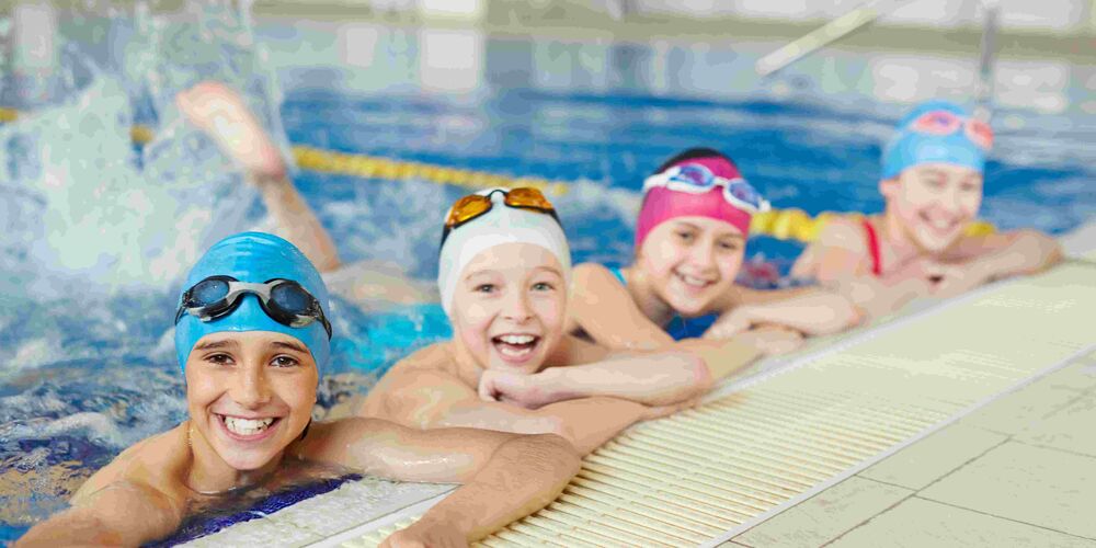 Vier Kinder mit Badekappen sind im Wasser vom Hallenbad und halten sich lächelnd am Beckenrand fest.